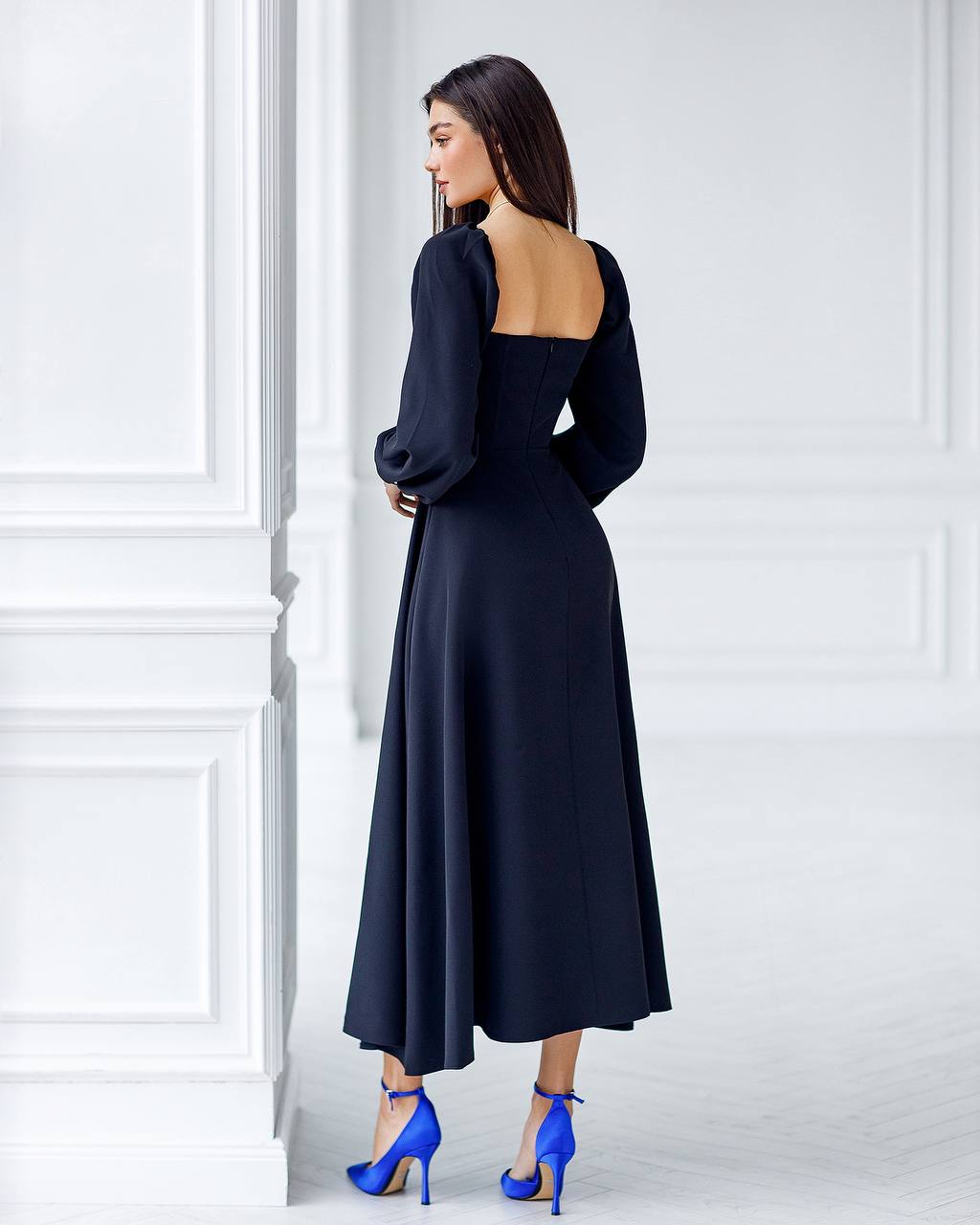 Woman wearing a black dress with long sleeves and open back, standing against a white wall.