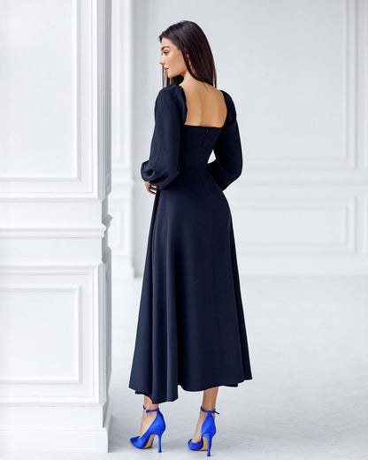 Woman wearing a black dress with long sleeves and open back, standing against a white wall.