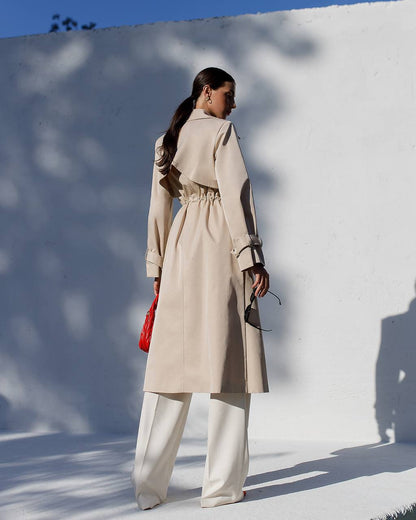 Woman in a beige trench coat standing against a white wall with blue sky.