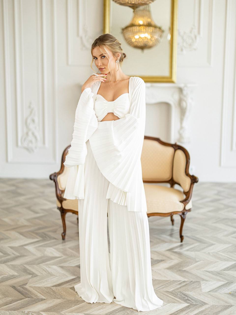 A woman models a white bohemian-style two-piece outfit consisting of a cutout sweetheart top with long draped sleeves and matching pleated wide-leg pants, posed in an elegant room with a vintage sofa and herringbone wood floor.
