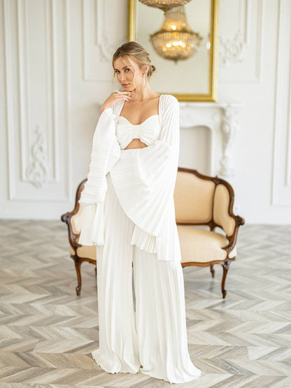 A woman models a white bohemian-style two-piece outfit consisting of a cutout sweetheart top with long draped sleeves and matching pleated wide-leg pants, posed in an elegant room with a vintage sofa and herringbone wood floor.