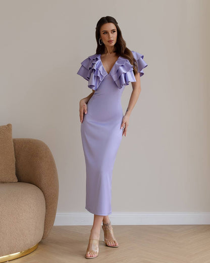 Woman wearing a lavender dress with ruffled shoulders in a room with a beige armchair.