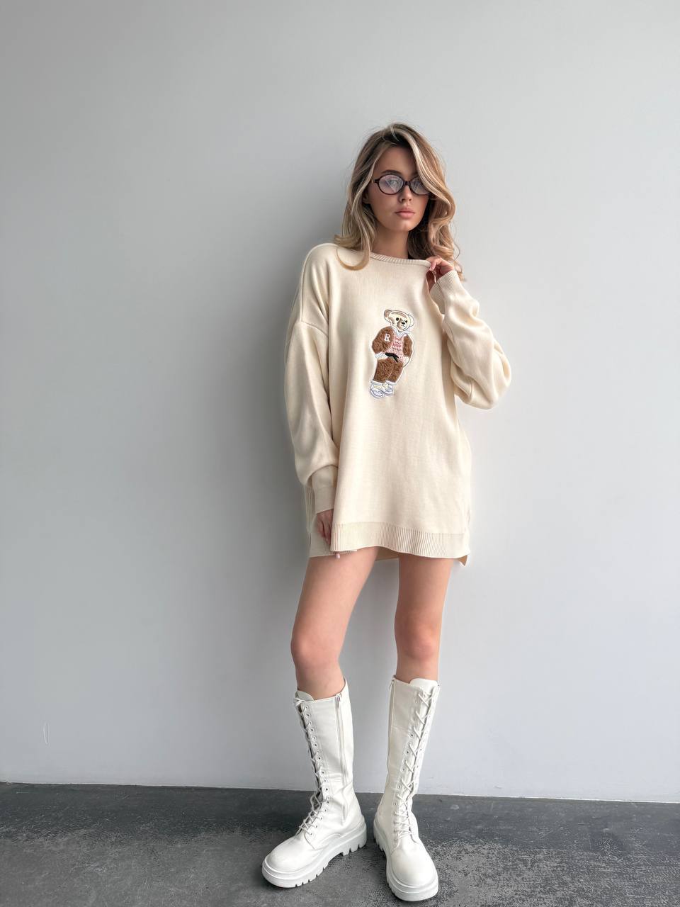 Woman wearing a beige sweater dress with a graphic design and white boots against a plain background