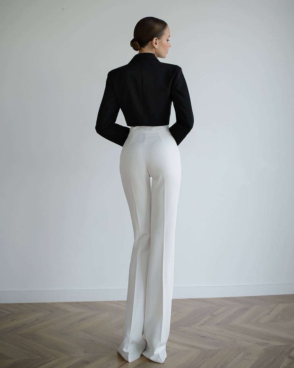 Person wearing a black top and white pants standing against a plain white wall.