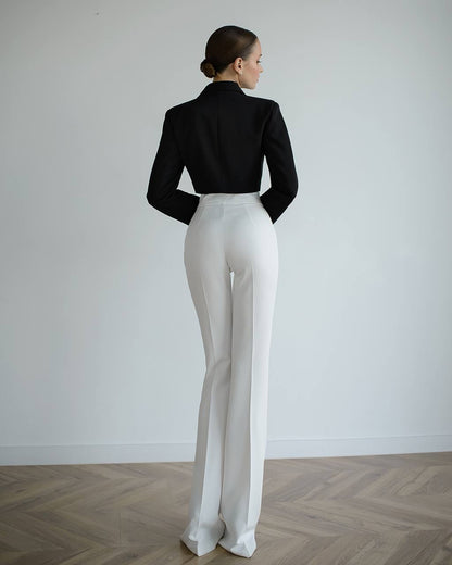Person wearing a black top and white pants standing against a plain white wall.