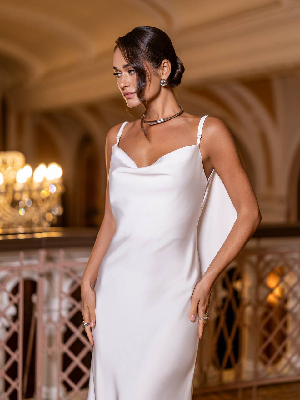 Woman in a white dress standing in an elegant indoor setting with chandeliers.