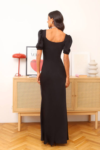 Woman in a black dress standing in a room with wooden flooring and a light-colored wall.