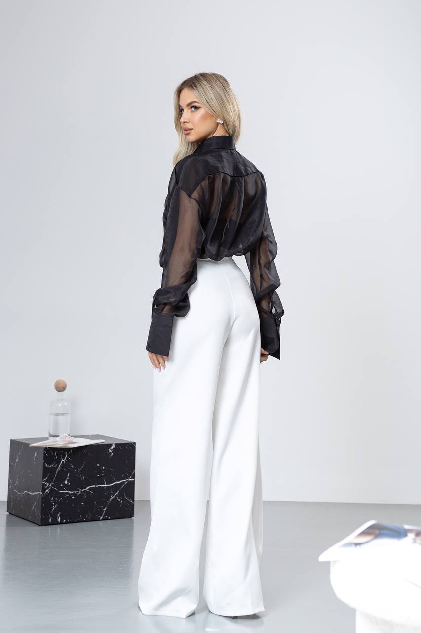 Woman wearing a black sheer blouse and white pants in a minimalistic setting