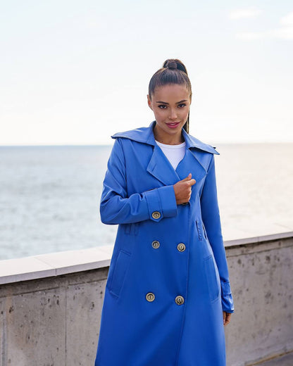 Woman wearing a blue coat with gold buttons by a waterfront.