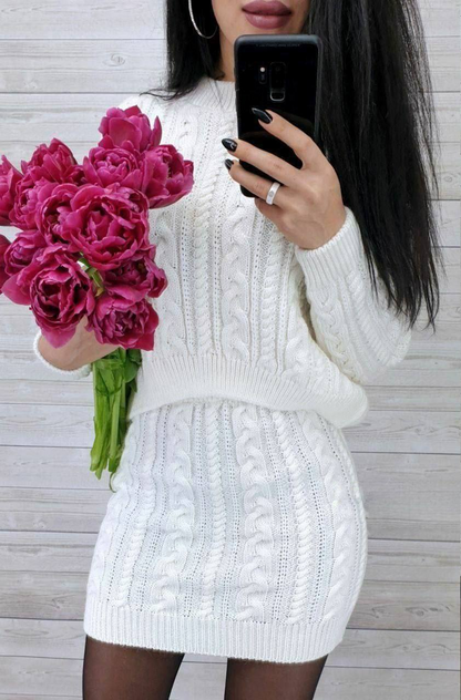 Person wearing a white cable knit dress holding pink flowers and a phone.