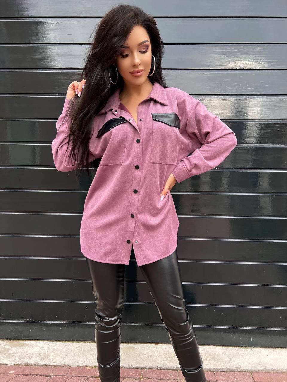 Woman wearing a pink shirt and black leather pants against a gray wall.