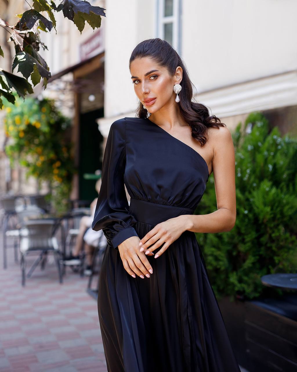 Woman in a black one-shoulder dress standing outdoors with greenery and a building in the background.