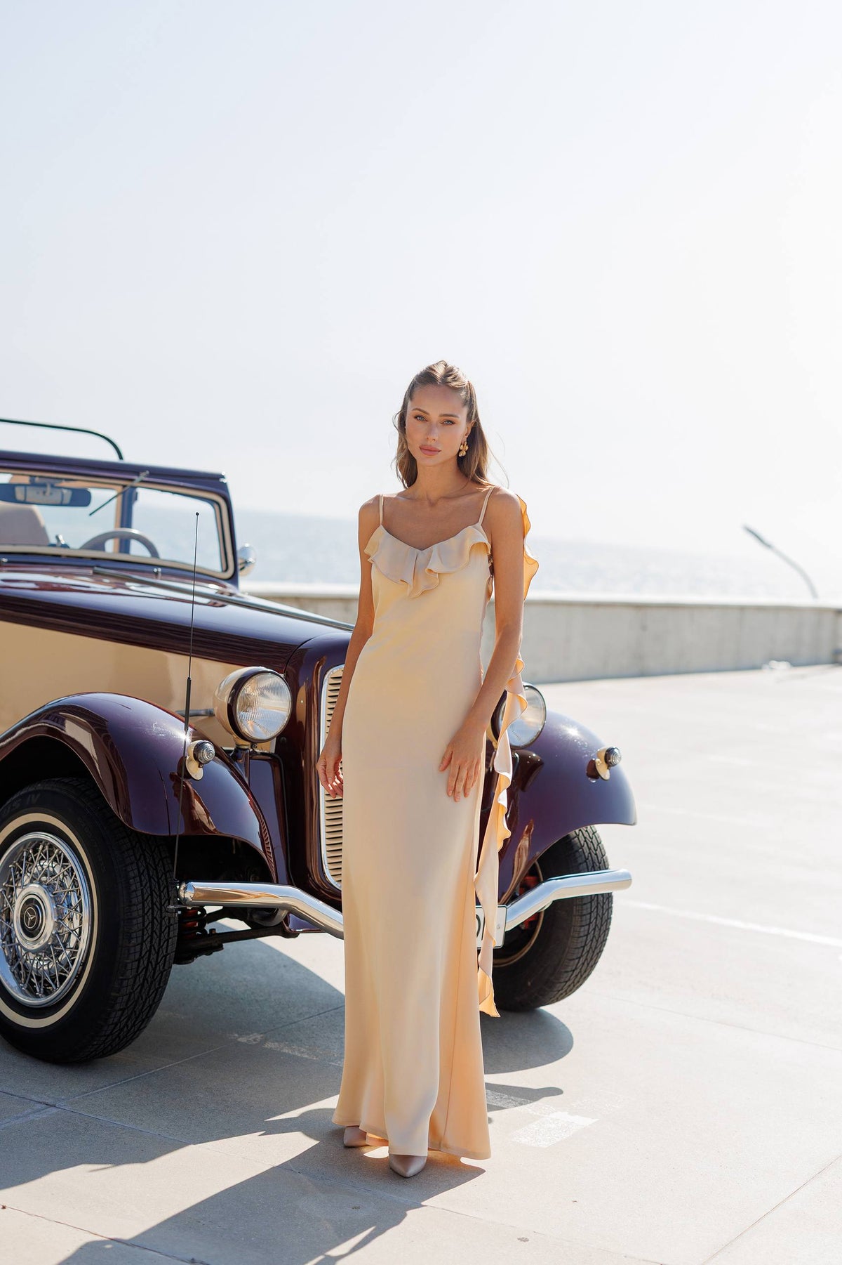 A woman wearing a floor-length cream satin slip dress with ruffled straps stands next to a vintage convertible car on a sunny outdoor paved area.