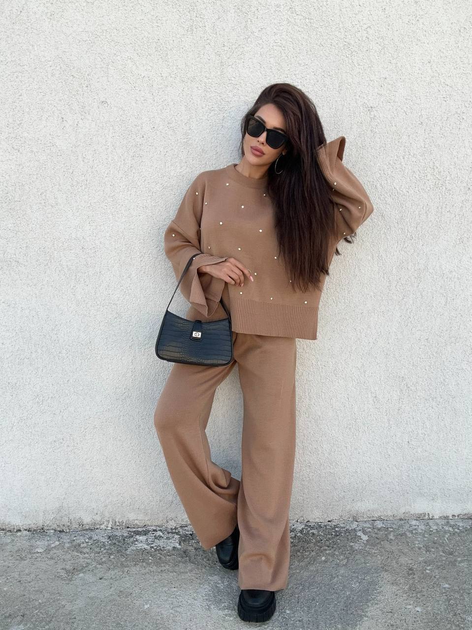 Woman in a beige outfit with a black handbag against a white wall