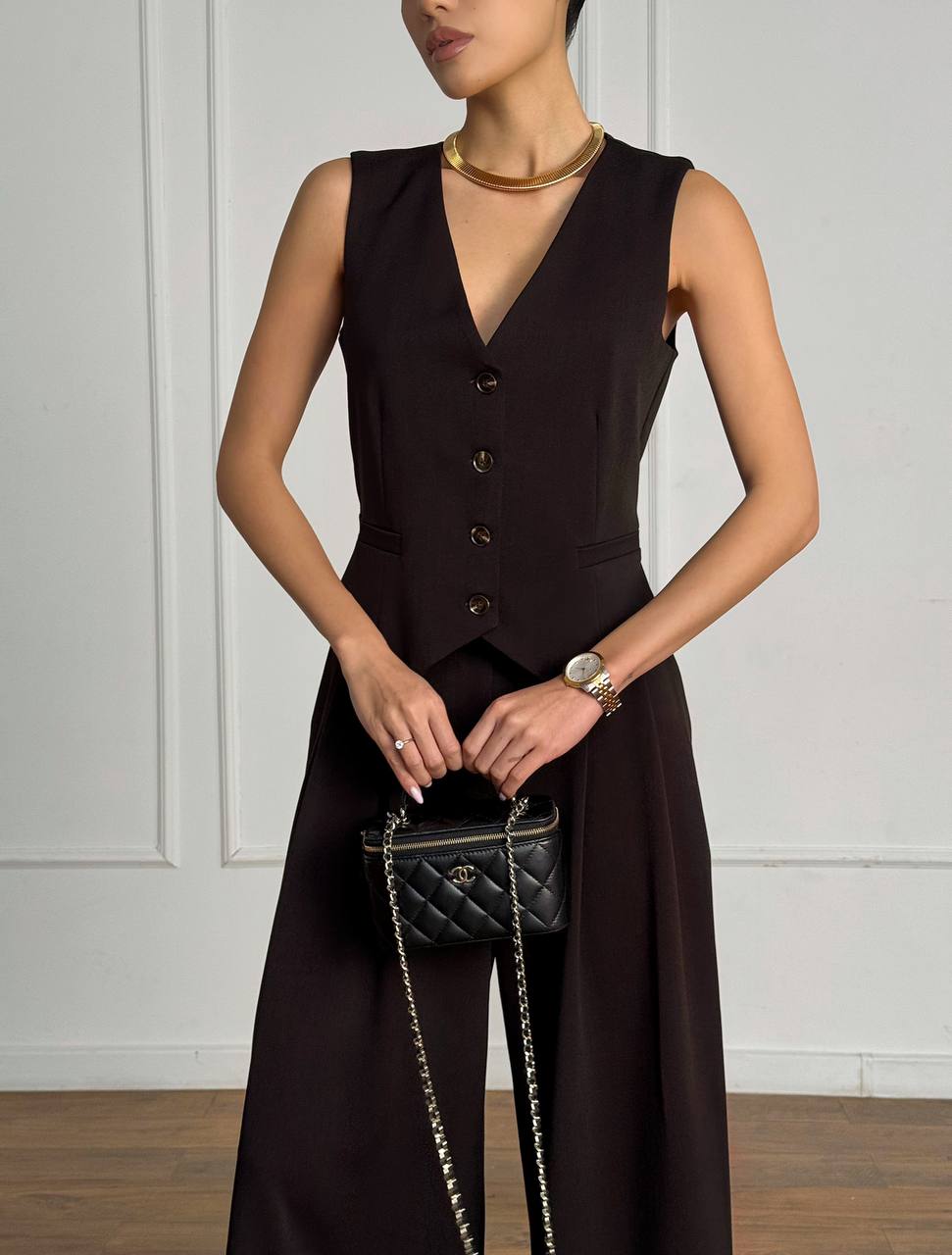 Woman wearing a dark brown sleeveless top and long skirt holding a black handbag against a white wall.