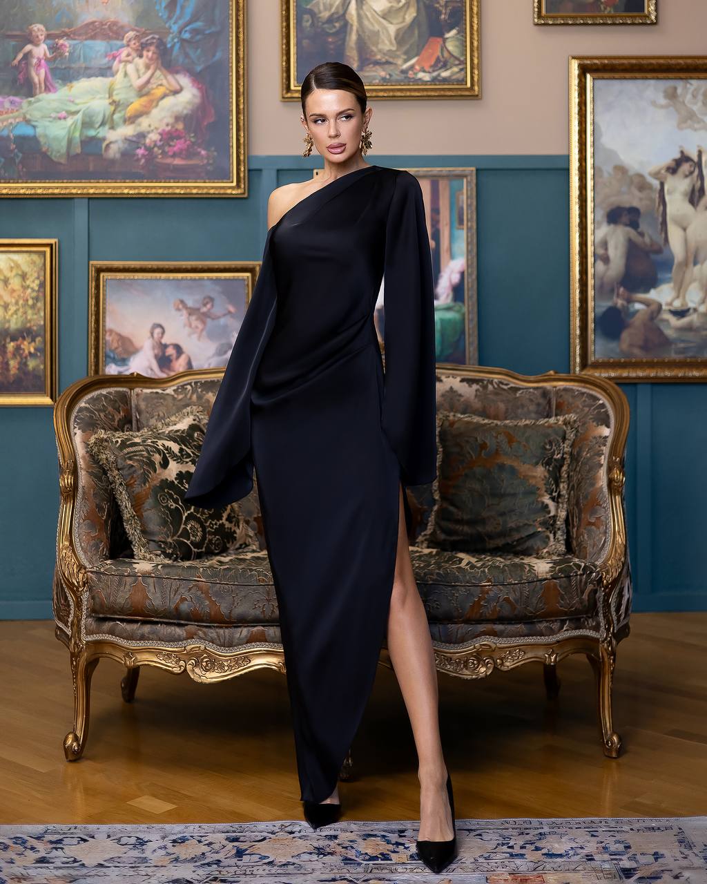 A woman models a floor-length, one-shoulder black gown with a high thigh slit, standing in a vintage room with an ornate sofa and framed paintings on the wall.