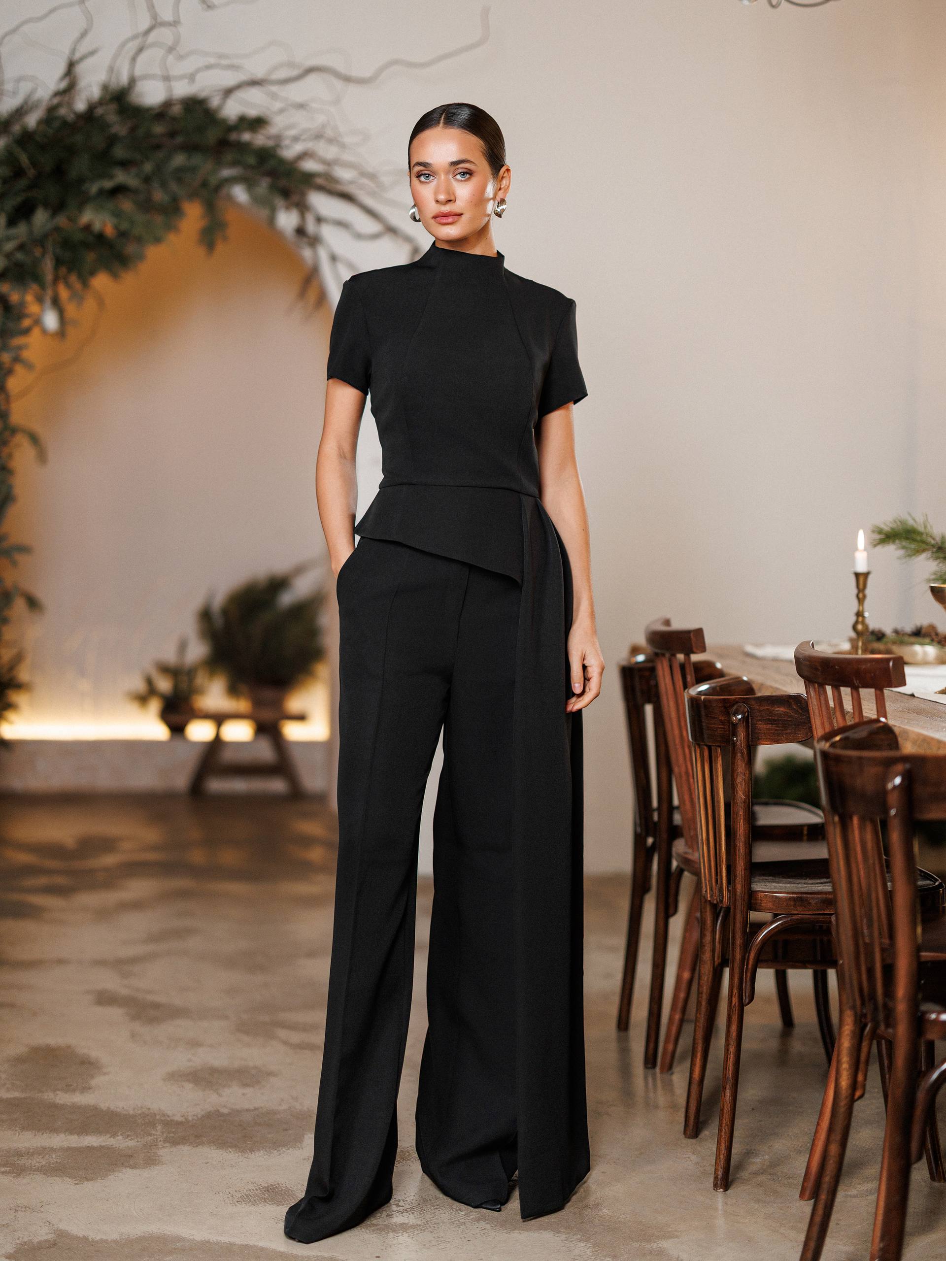 Woman in a black outfit standing in a room with wooden furniture and plants.