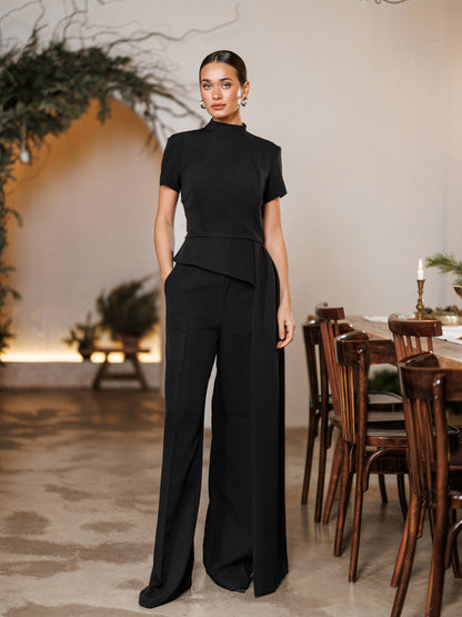 Woman in a black outfit standing in a room with wooden furniture and plants.