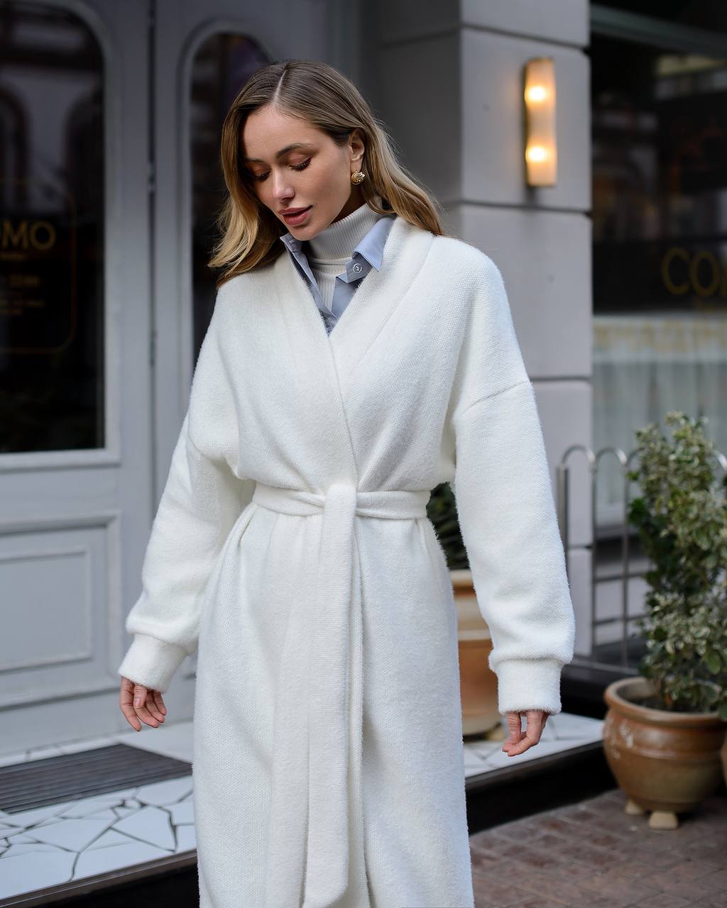 Woman wearing a white robe standing outside a building with a plant in the foreground.