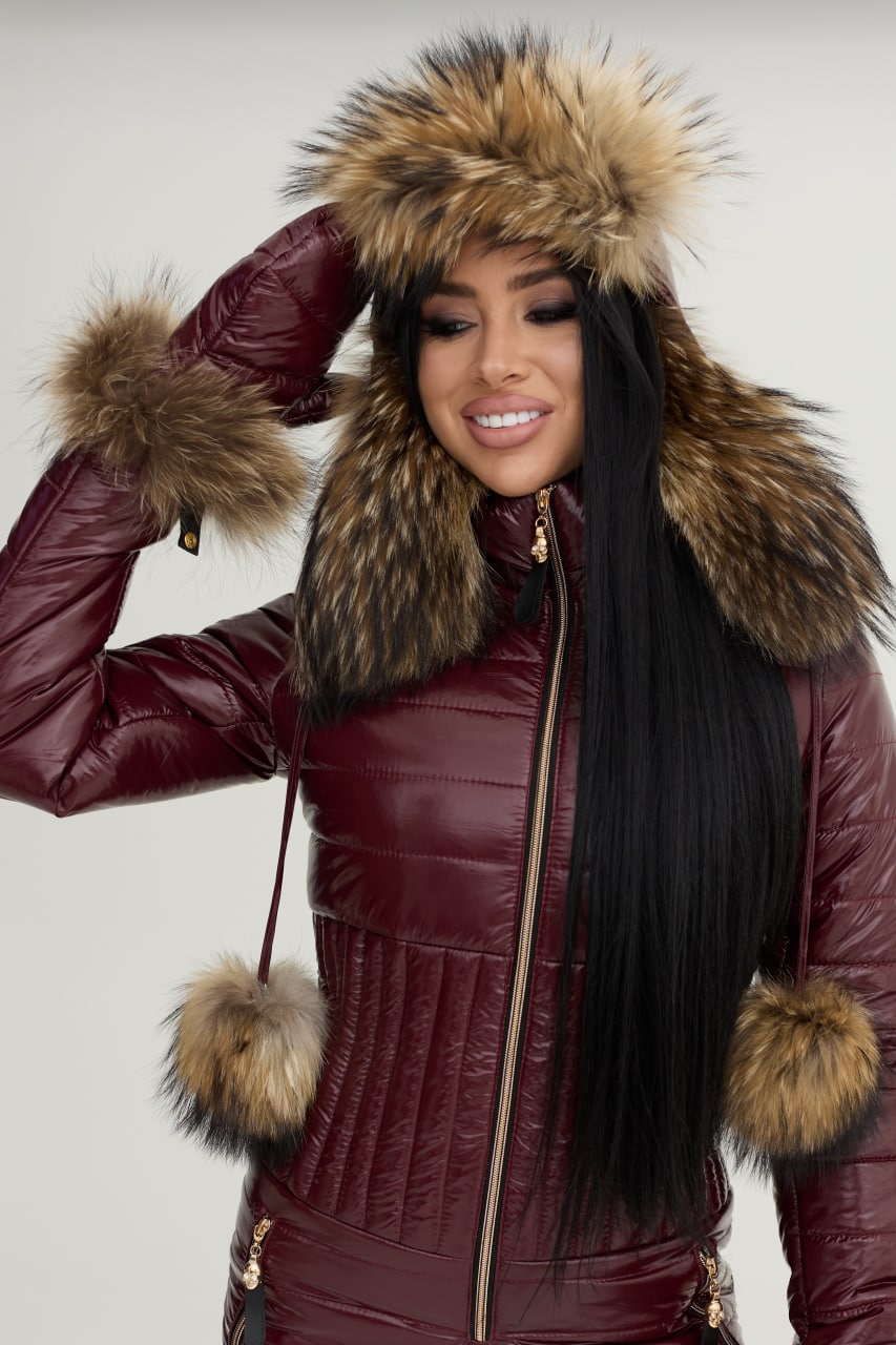 Woman wearing a burgundy puffer jacket with fur-lined hood and cuffs on a plain background