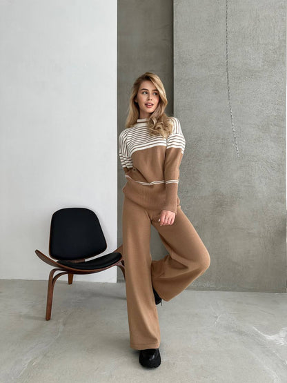 Woman in a striped sweater and brown pants standing against a concrete wall with a black chair.