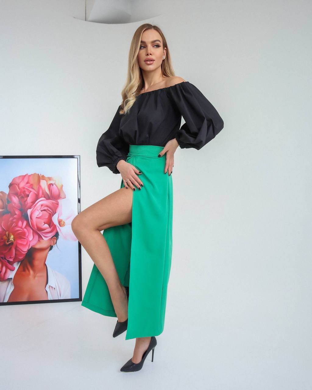 Woman wearing a black off-shoulder top and green skirt posing in a room with a decorative headpiece on the wall.