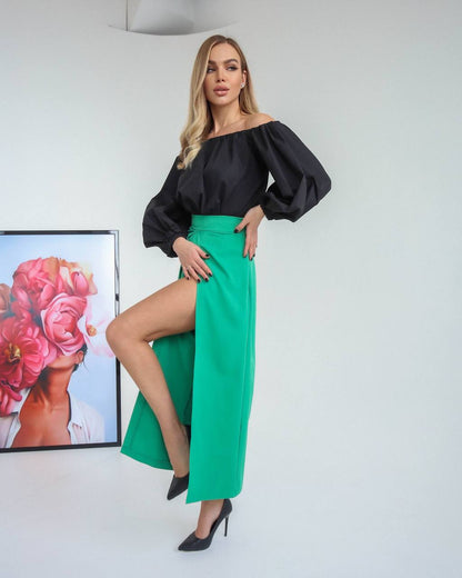 Woman wearing a black off-shoulder top and green skirt posing in a room with a decorative headpiece on the wall.