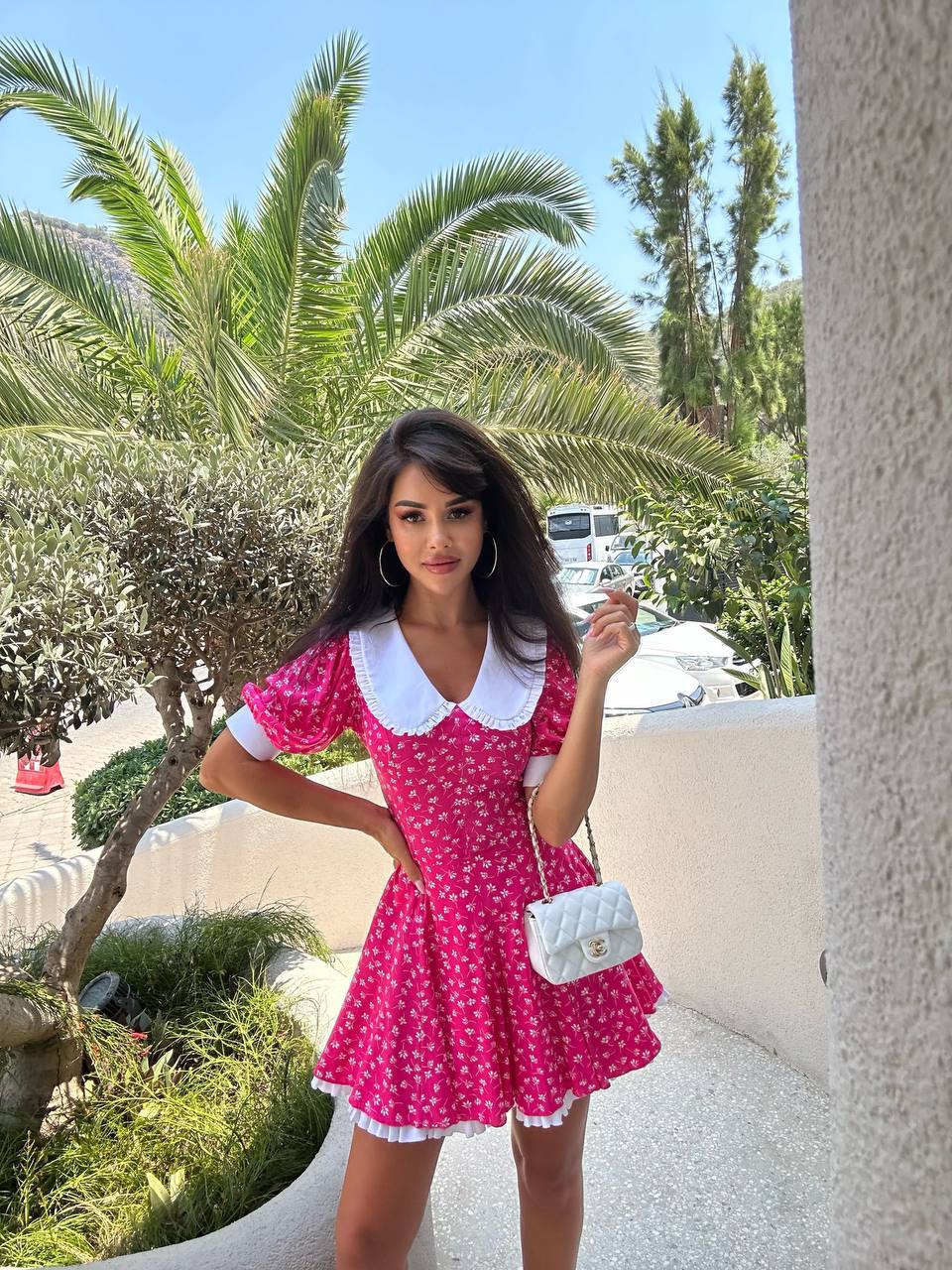 Woman in a pink dress with white collar and trim standing outdoors with palm trees in the background.