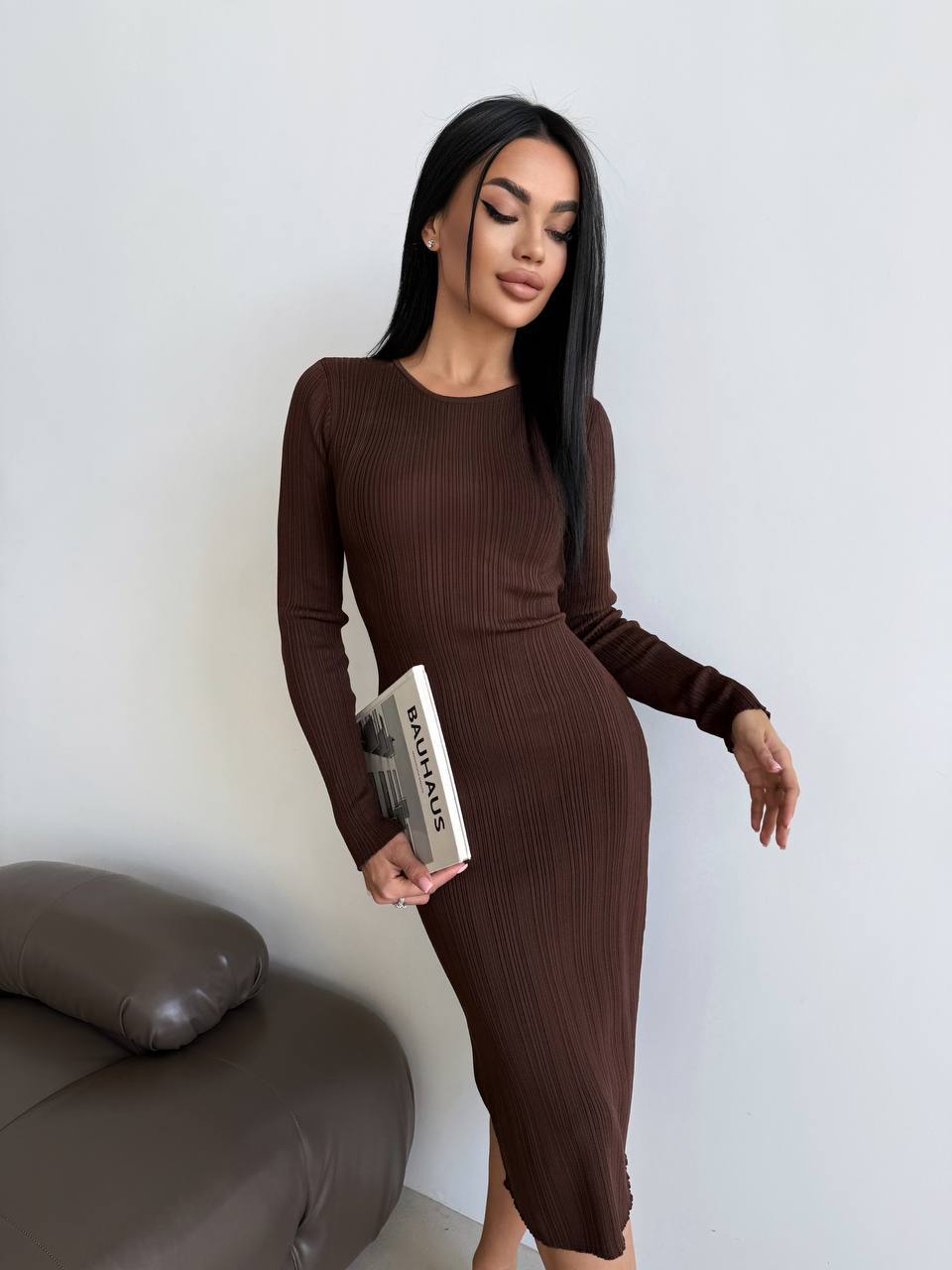 Woman in a brown ribbed dress holding a book against a white wall.