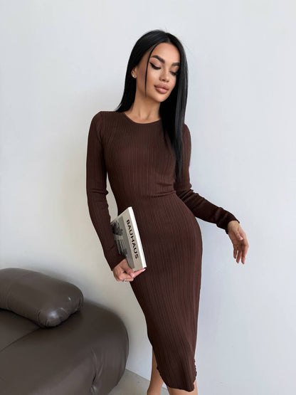 Woman in a brown ribbed dress holding a book against a white wall.