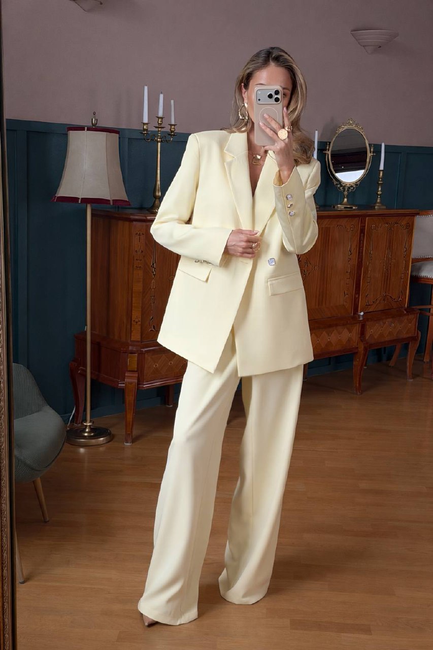 Person wearing a light-colored suit taking a mirror selfie in a room with wooden furniture and a lamp.