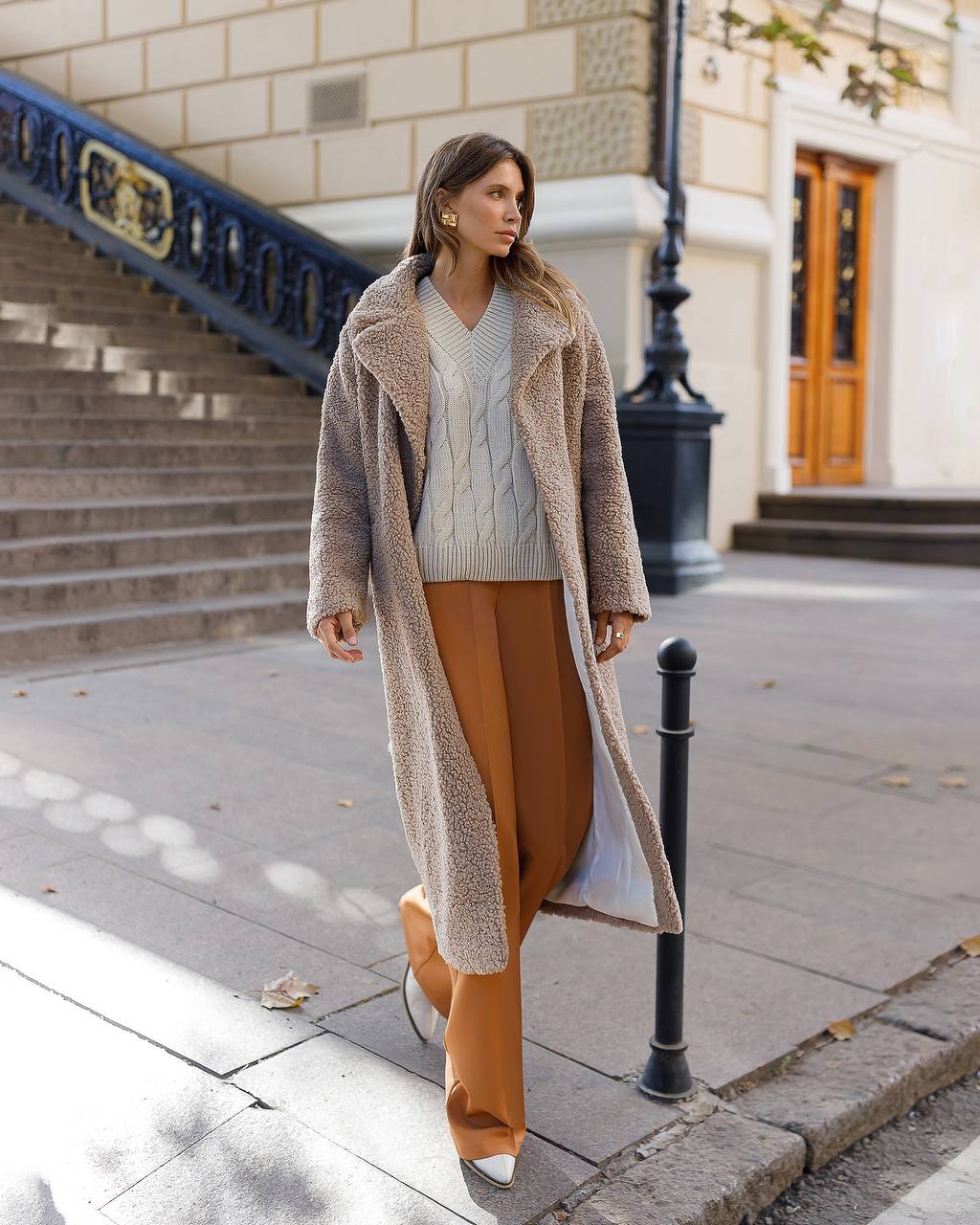 Woman walking on a sidewalk wearing a long coat, sweater, and pants.