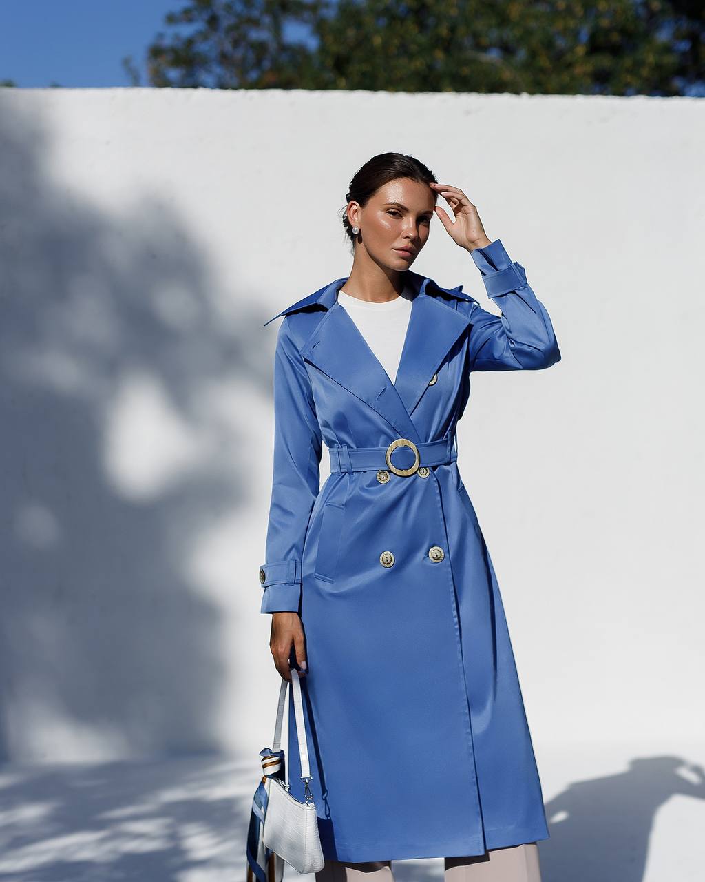 Woman wearing a blue trench coat against a white wall with shadows.