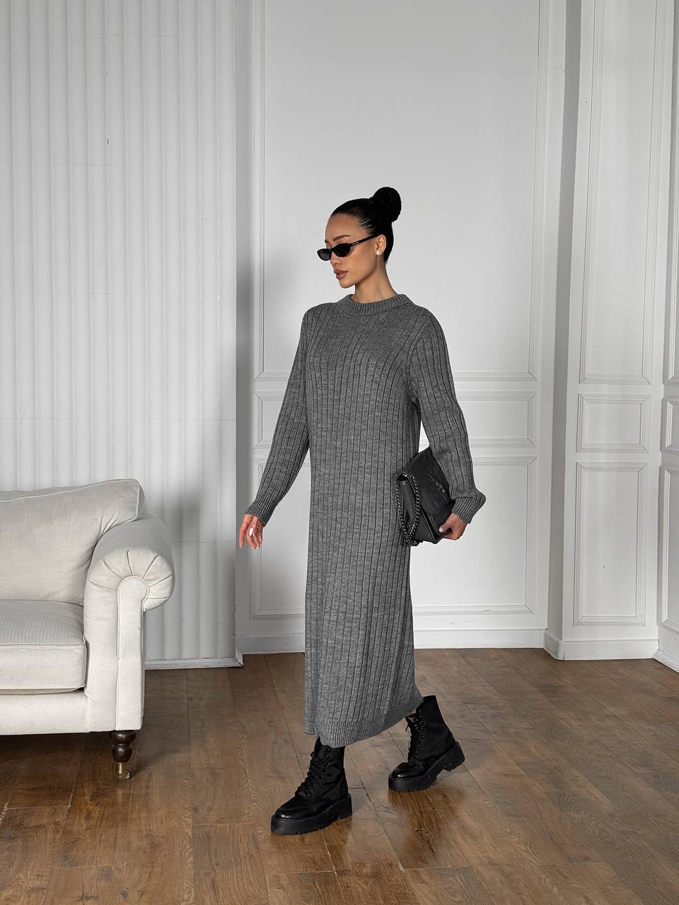Woman in a gray knit dress standing in a room with white walls and a gray sofa.