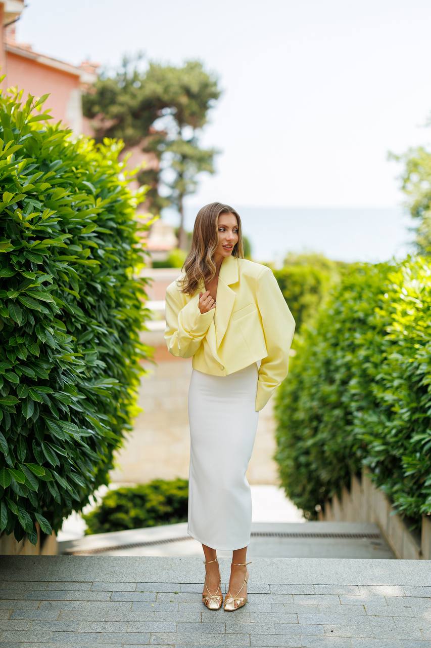Woman in a yellow jacket and white skirt standing in a garden with greenery and a building in the background.