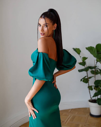Woman in a green off-shoulder dress posing indoors with a plant in the background