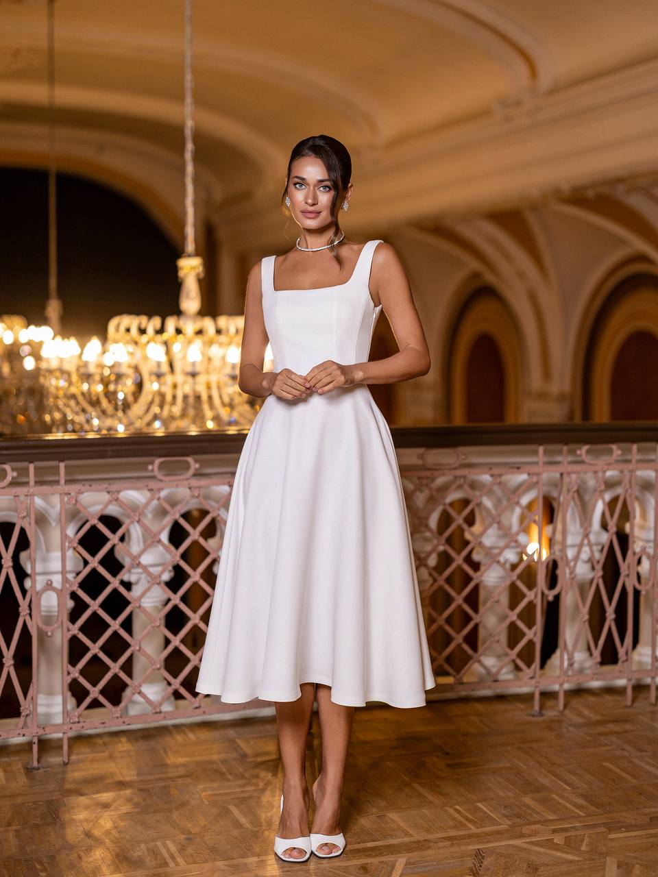 Woman in a white dress standing in an elegant indoor setting with chandeliers.