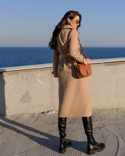 Woman in a beige coat and black boots standing by the ocean