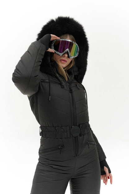 Person wearing a black snowsuit with a fur-lined hood, holding colorful goggles.