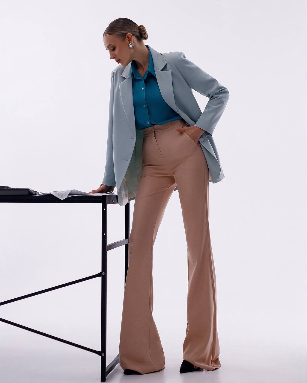 A woman models a light blue blazer over a teal button-up shirt and beige flared trousers, standing beside a black table against a clean white studio background.