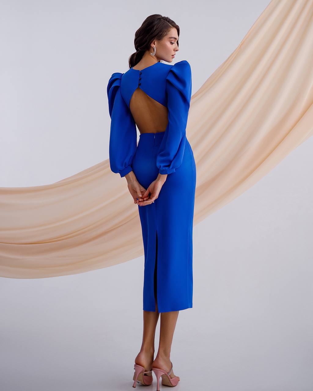 Woman wearing a blue dress with a backless design, standing against a beige curtain.