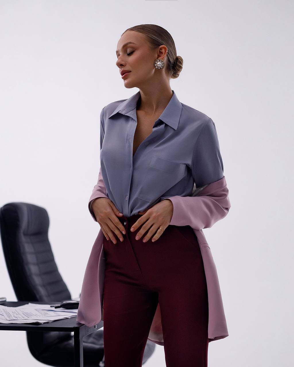 A woman models a color-blocked long-sleeve button-up shirt with a slate blue upper body and lavender sleeves, paired with burgundy trousers, in a minimalist studio setting.