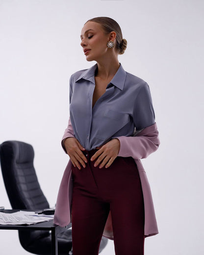 A woman models a color-blocked long-sleeve button-up shirt with a slate blue upper body and lavender sleeves, paired with burgundy trousers, in a minimalist studio setting.
