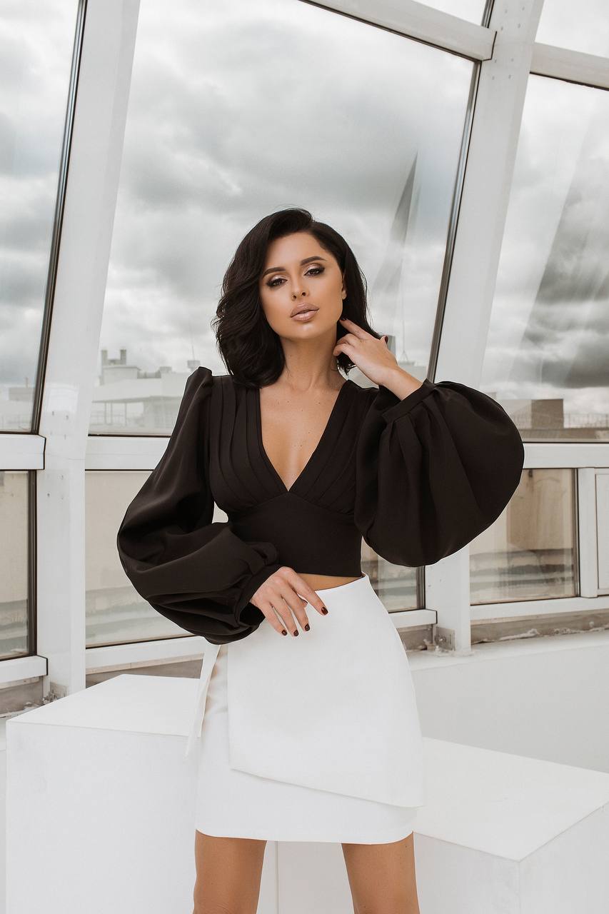 Woman wearing a black top and white skirt in front of large windows with a cityscape view.