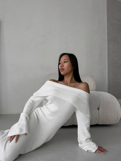Woman wearing a white off-shoulder sweater and pants sitting on a gray floor against a plain wall.