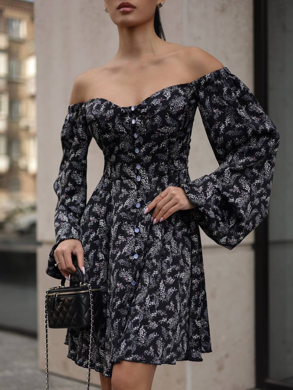 Woman wearing a black floral off-shoulder dress with long sleeves, standing against an urban background.