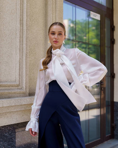 Woman wearing a white blouse with a bow tie and dark pants standing in front of a building entrance.