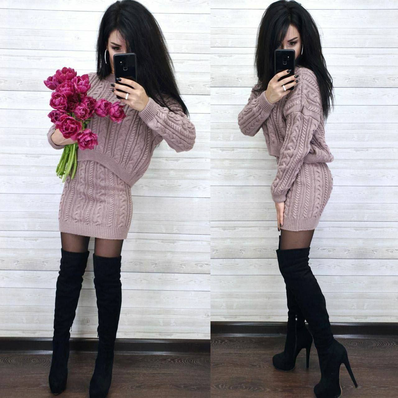 Woman in a pink cable knit dress with black knee-high boots holding pink flowers, taking a mirror selfie.