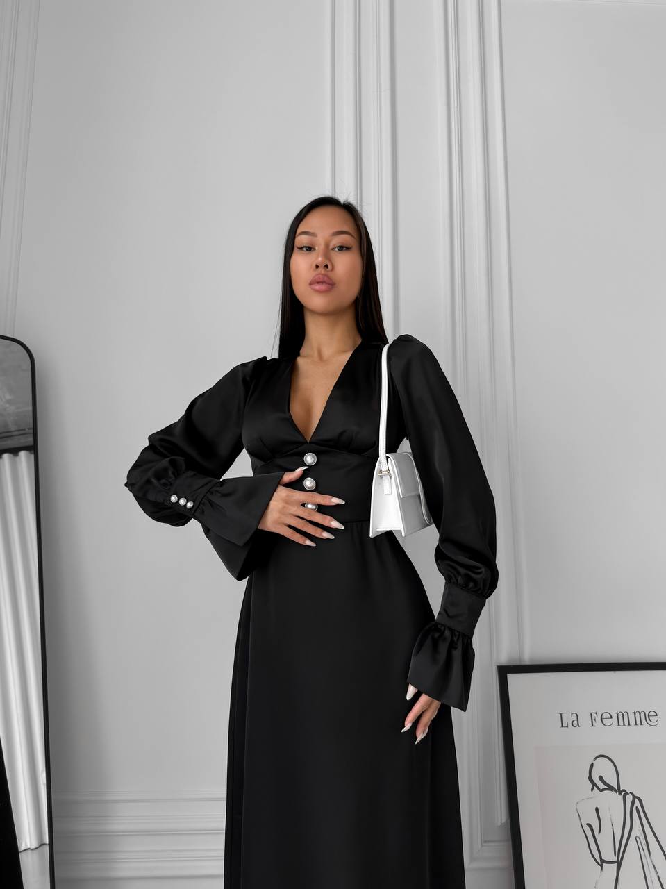 Woman wearing a black dress with a white bag against a gray wall.