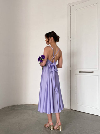 Woman in a lavender dress holding flowers against a plain wall.